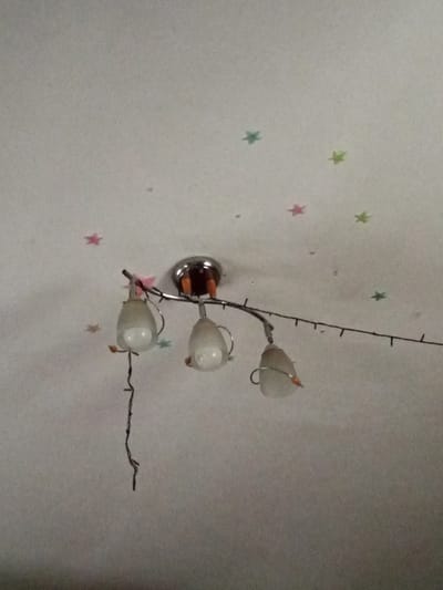 The image shows a ceiling light fixture with three hanging lamps. The ceiling is white and appears to have several small, star-shaped stickers in various colors, including pink, green, and blue. The light fixture itself has a silver-colored arm extending from a central mount, and the lamps are encased in frosted glass with silver accents. A thin, dark wire hangs from the fixture, separate from the lamp shades. The lighting in the image is somewhat dim, suggesting it may be nighttime or the lights are off. The image does not provide any information that would allow for its location to be determined within the city of Shostka, Ukraine.
