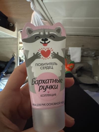 A close-up, slightly upward-angled image depicts a person's left hand holding a tube of hand cream within a train compartment. The hand cream tube is predominantly white and pink, featuring a cartoon raccoon character at the top, depicted with a happy expression and holding a glowing red heart. Russian text on the tube includes "ПОХИТИТЕЛЬ СЕРДЕЦ" (Heart Thief), "Бархатные ручки" (Velvet Hands), "КОЛЛЕКЦИЯ" (Collection), and "КРЕМ ДЛЯ РУК ОСНОВНОЙ УХОД" (Hand Cream Basic Care). The hand is fair-skinned, with visible fingernails, gripping the lower portion of the tube.
The background indicates an interior train setting. To the left, a metallic frame consistent with a bunk bed structure is visible, alongside a blue strap. To the right, a person's lower leg is present, clad in dark trousers and a brightly colored orange sock with green patterns. A white, folded towel hangs on a fixture in the upper right. Other blurred fabric items are discernible in the background. The interior walls and ceiling are light-colored. This scene is set within a train compartment, consistent with travel in Russia, potentially in the vicinity of Gryazi.