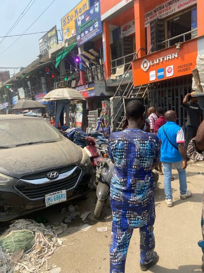 This image depicts a bustling commercial street scene in Igbede, Nigeria, under a hazy daylight sky. In the immediate foreground, a man wearing a blue and white patterned traditional Nigerian tunic and trousers stands with his back to the camera, facing the right side of the frame. To his right and slightly behind him, another man, dressed in a light blue short-sleeved shirt, light blue jeans, and white sneakers, stands facing the same direction, carrying a black shoulder bag.
To the left, a dark-colored, dust-covered Hyundai SUV is parked, displaying a Nigerian license plate "UMG-686RU". Behind the SUV, a maroon and black scooter is partially visible. Between these vehicles and extending into the mid-ground are substantial piles of folded denim jeans, arranged on what appears to be a vendor's stall. An open, light-colored umbrella is propped above these goods, likely providing shade or serving as a display element. A closed black umbrella is also visible in this area.
The background is dominated by multi-story commercial buildings that line the street. Various signs are visible on these structures, including "SSU-KOM," "VENUSOLAR," "INDEX BATTERIES," and "LOBAL NIGERIA LIMITED." The building on the far right is orange and features a prominent sign for "OLUTION" (Lutian), along with an external metal staircase. Numerous electrical wires crisscross overhead. The ground consists of a dusty, uneven surface, littered with various debris including plastic bottles, wrappers, and fabric scraps. Several other individuals are present in the mid-ground and background, either standing or walking, contributing to the active street environment.