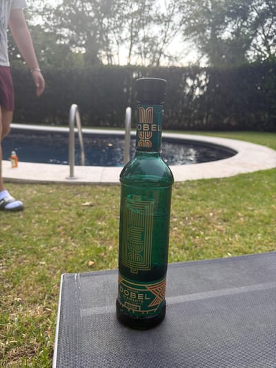 A green bottle of Dobel Tequila Diamante lies horizontally on a gray, mesh-covered surface, likely part of an outdoor chair or lounger. The bottle is positioned with its label facing upward. The surrounding environment suggests an outdoor setting, possibly a backyard or patio. In the background, there is a swimming pool with a curved edge and visible pool ladders. Green grass is visible on either side of the pool. A person is visible in the bottom left, wearing a white shirt and burgundy shorts. A foot wearing a white sock and blue-and-white shoe is visible near the bottom center. The image appears to be taken outdoors on a sunny day. The location is near a swimming pool, which suggests a residential or recreational area. Santa Ana Yancuitlalpan (Chilhuacán), Mexico cannot be determined from the available visual evidence.