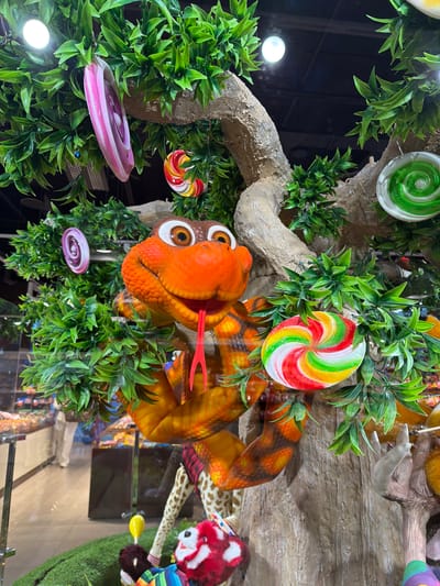 The image captures a whimsical display, likely at a confectionery or a themed attraction, in Sokilnyky, Ukraine. A prominent feature is a tree-like structure adorned with artificial green leaves. Hanging from the branches are several large, colorful, swirl-patterned lollipops, resembling those found in candy shops. These lollipops are pink and white, red, yellow, green, and white, and green.
A central focus is an animated, orange snake sculpture with large, wide eyes and a protruding red tongue. The snake's body is coiled around the tree trunk and branches, its scales detailed with patterns of orange, brown, and yellow.
In the background, through a glass pane, a store interior is visible. Shelves stocked with various candies are apparent, along with a person in casual attire walking in the distance, suggesting a retail environment. The foreground also shows portions of other plush toy-like decorations on a green, grassy base, hinting at a playful and festive setting. The lighting suggests an indoor environment, possibly a well-lit shop or exhibition space.
