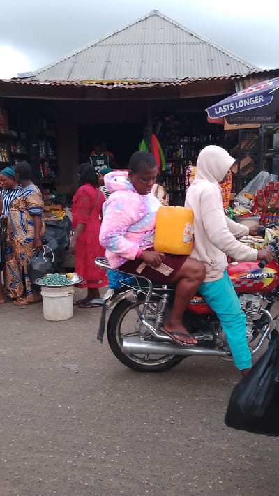 Two individuals are riding on a red motorcycle on a paved road. The person in the back, wearing a multi-colored hooded jacket and a maroon skirt, is holding a large yellow jerrycan. The person in front, wearing a light-colored hooded sweatshirt and teal pants, is driving the motorcycle. A black plastic bag is visible in the foreground on the right. In the background, there is a market stall with a corrugated metal roof, displaying various items for sale. Several other people are visible in the background, some browsing the stalls and others walking. The overall scene depicts a busy market environment in Agbarho, Nigeria.