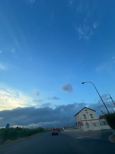 The image depicts a street scene in Manfredonia, Italy, near the intersection of Via Pietro Nenni and Via Luigi Capuano, at dusk. The sky is a deep blue with scattered clouds, transitioning from light to dark as it approaches the horizon. On the right side of the road, a white building with red-trimmed windows and doors stands prominently. A street sign pointing to "Gargano" and "Riponti" is visible in front of the building. A lone street lamp with a curved arm extends over the road. In the foreground, a dark asphalt road curves to the left, with a silver car in the lane ahead. To the left of the road, there is a dense green hedge or vegetation, and a blue sign with a white "P" symbol for parking is visible. In the distance, faint lights suggest other buildings or structures, and power lines are visible against the sky. The overall atmosphere is calm and transitional, suggesting the end of the day.