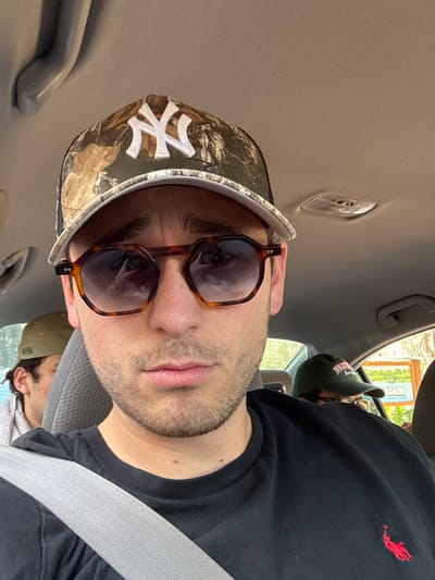 An eye-level photograph depicts a male subject seated in the front passenger seat of a vehicle, located in Micanopy, United States. The primary subject wears a camouflage baseball cap with a white New York Yankees logo, octagonal tortoiseshell-patterned sunglasses with gradient lenses, and a black short-sleeved t-shirt featuring a red Ralph Lauren polo player emblem. A gray seatbelt is visible across his chest. His facial expression is neutral. Behind the main subject, another male individual wearing an olive-colored baseball cap is partially visible on the left. Further back and to the right, a third person is partially seen, wearing a dark green baseball cap and glasses. The vehicle's interior has a light-colored headliner and gray fabric seats. Through a rear window on the right, an outdoor sign with a white background and multi-colored elements is visible, displaying the partial text "...AGAIN!".
