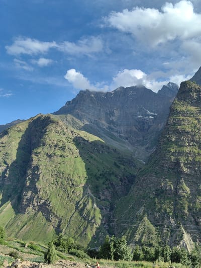 The image depicts a mountain landscape in Tiling, India, under a bright blue sky dotted with fluffy white clouds. The foreground features terraced fields with green vegetation and scattered trees, suggesting agricultural activity. The middle ground is dominated by steep, green mountainsides, carved with visible contours and shadows, indicating the uneven terrain. In the background, towering, rugged mountains rise, some with snow-capped peaks and scree slopes, showcasing the dramatic geological formations of the region. The overall scene conveys a sense of natural beauty and the raw power of the mountainous environment.