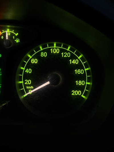 This is a close-up, dimly lit photograph of a car's dashboard, primarily featuring the speedometer and a fuel gauge. The dominant subject is the speedometer, a circular gauge with bright green markings and numbers illuminated against a dark background. The needle of the speedometer is pointing slightly past the 20 km/h mark, indicating the vehicle is moving at a slow speed. The numbers on the speedometer range from 0 to 200 km/h, with tick marks at 20 km/h intervals.
Below and to the left of the speedometer is a smaller, curved fuel gauge. Its needle is positioned between the "0" and "1/2" markings, suggesting the fuel tank is less than half full. The fuel symbol, a gas pump icon, is also visible near the gauge.
The overall scene is dark, suggesting it is either nighttime or the interior of a vehicle with its lights on. The green glow from the dashboard instruments is the primary light source in the image. The surface of the dashboard appears slightly dusty or textured. There are no people visible in the image. The location context provided suggests this might be the dashboard of a car in Emelyanovo (airport), Russia, though this cannot be confirmed solely from the visual information within the image itself. The visible text includes numerical speed markings (0, 20, 40, 60, 80, 100, 120, 140, 160, 180, 200), the unit "km/h", and markings for the fuel gauge ("0", "1/2", "1").