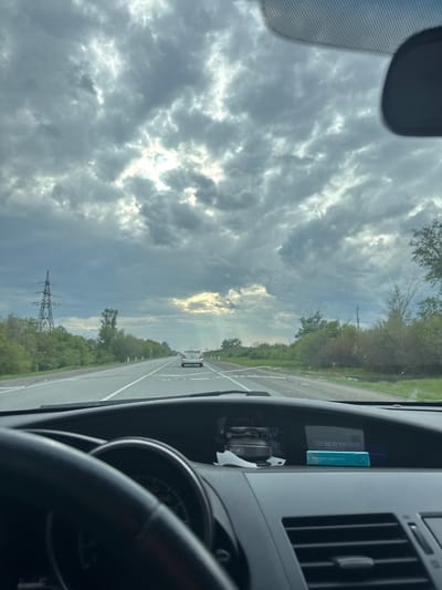 The image depicts a view from inside a car driving on a highway. The sky is filled with dark, ominous clouds with a patch of sunlight breaking through. The road stretches into the distance with trees lining either side. A power line tower is visible in the distance. The dashboard of the car is visible, including the steering wheel, dashboard vents, and a blue package labeled "Music." Another car can be seen on the road ahead. The scene does not provide any information to confirm the location is Petrovka, Russia.