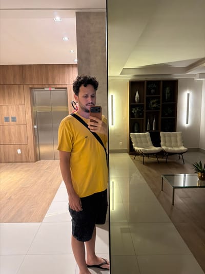 A man in a yellow shirt and black shorts stands in front of a large mirror, taking a selfie. The mirror reflects an apartment lobby with chairs, a decorative shelf, and tile flooring. Behind the man is a wooden-paneled wall with an elevator and recessed lighting. He holds a phone in his right hand, his head slightly tilted. The photo is taken indoors, showcasing a contemporary and clean interior design.