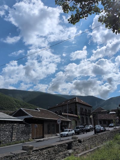 The image depicts a street scene in Şəki, Azerbaijan, likely during daytime, under a partly cloudy sky. Dominating the scene are several buildings, one with a distinctive arched facade, another with a modern, angular extension. The buildings are constructed with stone and brick, featuring tiled roofs. A street runs alongside the buildings, lined with parked cars. Among the vehicles, a white sedan is prominently visible in the foreground, and several other cars are parked along the street. Some people are visible near the white car. A speed limit sign indicating "40" is mounted on a pole. The street is bordered by a stone wall and a strip of green grass. In the background, a verdant, tree-covered mountain slopes upwards, suggesting a mountainous or hilly geographical setting. The overall impression is one of a settled, possibly historical, urban or semi-urban environment nestled within a natural landscape.