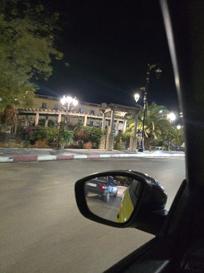 A car's side mirror reflects a vehicle at night on a road next to a building with illuminated ornate lights. Palm trees and other foliage are visible beside the structure. The road is striped with a red and white curb. The sky is dark, contrasting with the bright artificial lights and the car's headlights reflected in the mirror. A portion of the car's interior frame is visible on the periphery of the image.