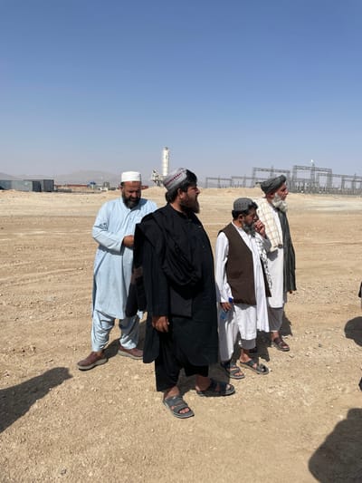 Four adult males are depicted standing outdoors on a dry, unpaved, light brown dirt or gravel surface under a clear, pale blue sky. The scene is located in Pir Dust, Afghanistan. All individuals are dressed in traditional attire: long tunics (shalwar kameez) and various head coverings.
From left to right:
1. A male in a light blue shalwar kameez and a white skullcap, with a dark beard, stands facing slightly right, hands clasped near his midsection. He wears brown shoes.
2. A male in a black shalwar kameez, with a black shawl draped over his left shoulder and a grey and red patterned cap, stands in the foreground facing right in profile. He has a dark, full beard and wears grey sandals.
3. A male in a white shalwar kameez, a brown sleeveless vest, and a dark grey patterned cap, stands facing right in profile. He has a shorter beard and holds a small blue plastic bottle in his left hand. He wears dark sandals.
4. A male in a white shalwar kameez and a dark patterned turban, with a long white beard, stands facing right in profile. He wears dark sandals.
All four individuals appear to be observing or listening to something or someone positioned to the right of the frame. Shadows of the men and an unseen object are cast on the ground, indicating bright sunlight. In the background, from left to right, are several shipping containers, followed by a tall white concrete batching plant with a "Z" logo, and further right, multiple unfinished metal lattice structures, consistent with a power substation or transmission infrastructure development. Distant, hazy mountains are visible under the clear sky.