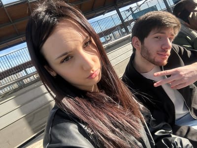 The image depicts three individuals outdoors under bright, direct sunlight, on what appears to be an elevated platform or walkway consistent with an urban transport station in Alfortville, France. The precise location within the city is not visually discernible.
In the immediate foreground, a young adult female with long, dark hair, dressed in a black leather jacket, poses with a slight pout and direct gaze into the camera. Sunlight casts distinct shadow lines across her forehead and right cheek.
To her right and slightly behind, a young adult male with light brown hair and a short beard wears a black jacket over a white ribbed garment. He holds his right hand in a V-sign gesture and looks slightly to his right.
In the blurred background, a third individual, dark-skinned and wearing a dark jacket, is partially visible to the far right, appearing to be engaged with a mobile device held to their ear.
The environment includes overhead metallic structures and railings, with a clear blue sky visible above, indicative of an outdoor, elevated urban setting.