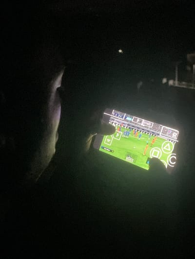 The image captures a person's hand holding a smartphone in a dark environment, likely at night or in a dimly lit room in Abatete, Nigeria. The smartphone screen is the primary light source, illuminating the hand and revealing that a video game is being played. The game displayed appears to be a soccer or football simulation, showing players in blue and orange uniforms on a green field.
The phone's interface clearly shows virtual controls typical of a PlayStation Portable (PSP) emulator, with buttons like 'Triangle', 'Circle', 'Square', 'X', and a directional pad (D-pad) on the left. Text visible on the screen includes "START," "SELECT," and "CITY FIVER," which might be a team name or a score indicator within the game. The player's thumb is visible near the bottom left of the screen, seemingly interacting with the virtual controls. Below the hand, a light-colored, indistinct object, possibly a pet or a piece of clothing, is partially visible in the dim light cast by the screen. The overall scene conveys a casual, private moment of gaming.