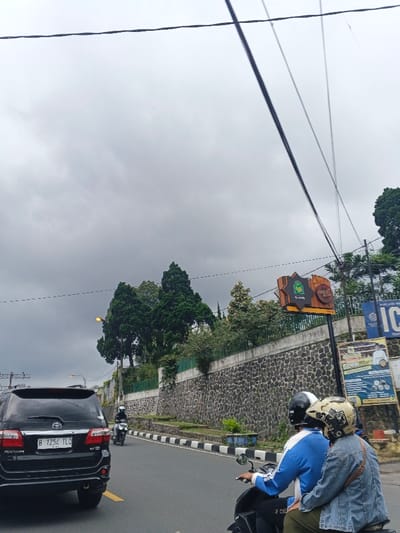 The image depicts a roadway scene under a heavily overcast sky. In the foreground, a black Toyota Fortuner SUV with Indonesian license plate "B 1252 TLO" proceeds down the road, with its left taillight illuminated. Immediately to the right of the camera's perspective, two individuals ride a dark-colored scooter; the driver wears a black helmet and a blue and white long-sleeved shirt, while the passenger wears a patterned helmet and a light blue denim-like jacket. Further ahead on the road, another person on a white scooter, wearing a white helmet and light jacket, is visible.
Along the right side of the road, a tall retaining wall constructed from dark, irregular stones supports an elevated terrain covered with dense green foliage, including several tall coniferous-like trees. Mounted on a pole along this wall is a brown, irregularly shaped commercial sign displaying "Cokelat Kita" (Indonesian for "Our Chocolate") in white text, alongside a green logo. Below this sign, other smaller informational posters are partially visible, including one blue sign with "ICE" text. Multiple black power lines crisscross the upper portion of the frame against the grey sky. Two streetlights with visible yellow-orange glows are present. The roadway itself is asphalt with white and black striped lane markings.
Based on the visible Indonesian license plate "B 1252 TLO" (indicating the Jakarta area) and the Indonesian language on the "Cokelat Kita" sign, the scene is located in Indonesia, not Palayan, Philippines.