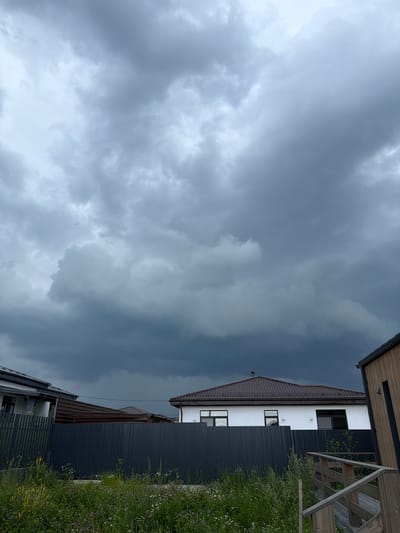 The image shows a suburban scene under a cloudy, stormy sky in Малое Седельниково, Russia. In the foreground, a wooden walkway leads up to a building on the right, with a dark metal fence and overgrown vegetation with wildflowers in front of it. Behind the fence are several houses, one with a white facade and a brown tiled roof, and another with a darker roofline. The sky is filled with heavy, dark gray clouds, suggesting an impending storm. A small, bright pink object, possibly a kite or balloon, is visible against the clouds in the upper left portion of the frame, between two houses.