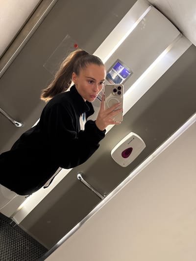 The image shows a young woman with a high ponytail taking a selfie in a mirror. She is wearing a black jacket and is holding a gold iPhone. In the background, there is a dispenser on the wall and a digital display. The environment appears to be an indoor public space, possibly a restroom or a train compartment. The lighting is bright and overhead, casting shadows. The location in the city of Harstad, Norway, is not discernible from the visual information in the image.