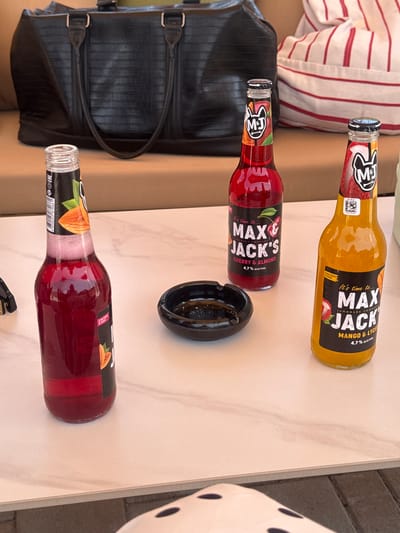 This media file shows a close-up, top-down view of three bottles of "Max & Jack's" lemonade on a light-colored, possibly marble-patterned surface. Two bottles are clearly visible, one with yellow liquid labeled "MANGO & LYCH" and the other with red liquid labeled "CHERRY & ALMOND". Both bottles have the "M&J" logo. A third bottle, also with red liquid, is partially visible at the bottom of the frame. Next to the red lemonade bottle is a black ashtray. To the left of the bottles, a black, textured bag with handles is visible. In the background, a person is wearing a white and red striped shirt. The lighting suggests it could be daytime, with natural light. The overall scene appears casual, possibly outdoors or in a relaxed indoor setting. The text "Max & Jack's", "MANGO & LYCH", "CHERRY & ALMOND", and "4,7% ALC/VOL" are legible on the bottles. The location context is Ptghni, Armenia.