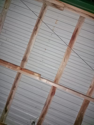 The image shows the underside of a corrugated metal roof supported by wooden beams. The beams are rough-hewn and show signs of age and weathering, with some discoloration and knots visible. A thin wire runs diagonally across the roof from the upper left to the lower right. The corrugated metal panels are a light gray color and are fastened with small nails or screws. The scene appears to be an interior shot, possibly of a shed, garage, or unfinished building, with no visible exterior elements or specific landmarks of Ақсай, Kazakhstan. There are no people or animals in the image. The lighting is dim, suggesting it might be nighttime or an overcast day.