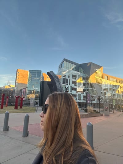 A woman with long, light brown hair and sunglasses stands on a sidewalk. Behind her are modern buildings, including one with a prominent sign reading "Passaro's". A large, abstract black sculpture is visible near the woman. Red vertical structures are lined up on a patch of grass. The sky is a clear blue. Metal bollards line the edge of the sidewalk.