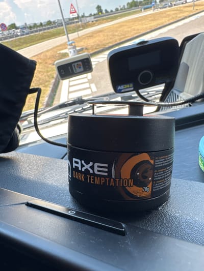 The image shows the interior of a vehicle, likely a truck, with a dashboard and windshield visible. On the dashboard, there is a black container of AXE Dark Temptation deodorant, weighing 70g. To the left of the deodorant is a black electronic device with a power symbol. In the background, through the windshield, a road and a grassy area are visible. There are no people in the image. The image does not provide any information about the location being in Occimiano, Italy.