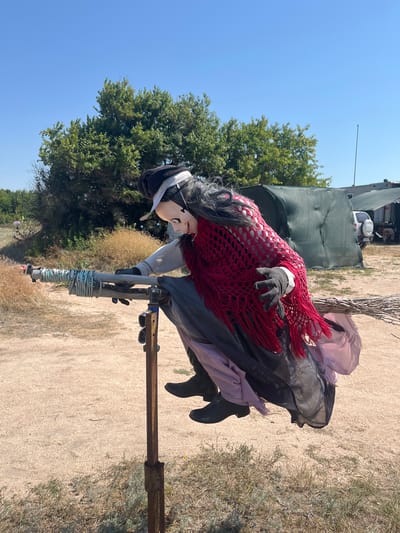 In Dolzhanskaya, Russia, a mannequin dressed as a witch is positioned outdoors, appearing to be riding a broomstick. The witch mannequin has long black hair, a pale mask-like face with a wide grin, a black hat, a white shirt, and a red crocheted shawl. It is wearing a long dark skirt and black boots. The broomstick is supported by a metal stand. The background shows a sandy ground, sparse vegetation, green trees, and some structures covered with tarps, under a clear blue sky.