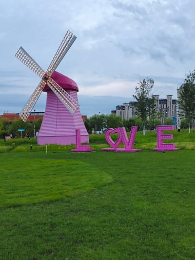 In Jiguan District, China, a pink windmill with illuminated blades stands prominently in a grassy park. To the right of the windmill, large pink letters spell out "LOVE" and "E", also illuminated with lights. In the background, modern apartment buildings are visible behind a line of trees. The sky is overcast with clouds. Several people are scattered throughout the park, some walking and others seated in the distance. The scene appears to be a public recreational area.