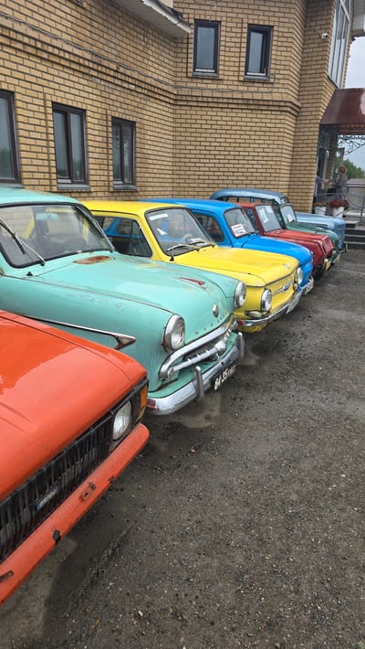 The image shows a line of vintage cars parked on a gravel surface next to a beige brick building. The cars are colorful, with a turquoise car in the foreground, followed by a yellow car, a blue car, and a red car further down the line. The building has several windows, and the ground appears wet, suggesting it might be raining or has recently rained. The sky is overcast, indicating a cloudy day. The scene is set in Bol'shoi Mashlyak, Russia. There is some visible text on the license plate of the turquoise car, which appears to read "BA 35-88 PB". No people are clearly visible in the image.