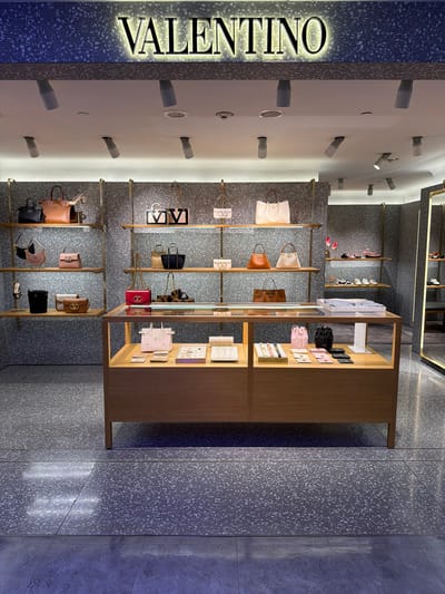 The image shows the interior of a Valentino store. The Valentino logo is prominently displayed on the left, illuminated with a golden glow. The store showcases handbags and accessories on shelves against a terrazzo-patterned wall. A wooden display case with various accessories stands to the right. Recessed spotlights illuminate the merchandise. The floor also has a terrazzo pattern. This scene is likely from the Valentino store in Taoyuan City, Taiwan.