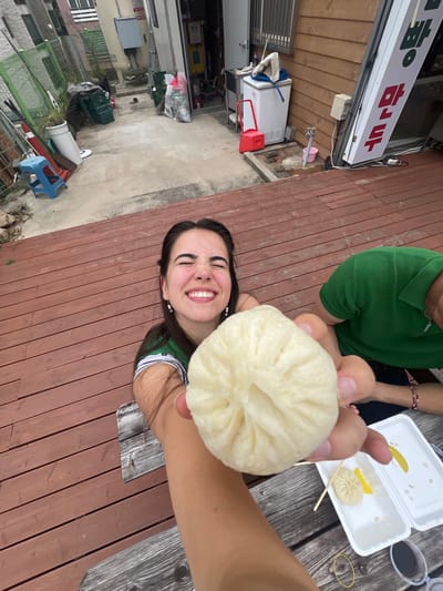 In Dongmyeong-myeon, South Korea, a young woman with dark hair and a green shirt grins widely as she holds a large, steamed bun towards the camera. The woman is on a wooden deck, and a person in a green shirt is seated to her right. In the background, a narrow alleyway is visible, with a fence on the left and a building with wooden siding on the right. The building has a sign with Korean characters, and a white refrigerator stands outside. The ground is concrete, with some scattered debris and plants.