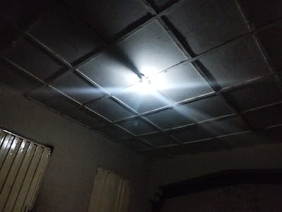 The image shows a ceiling with a grid pattern of square panels, illuminated by a single bright light source in the center. The light casts beams across the ceiling. The walls are painted a dark color, and there is a window with vertical slats on the left side of the image.  The location, Bajoga, Nigeria (latitude: 10.852, longitude: 11.426), is a small town in the northeastern part of the country.