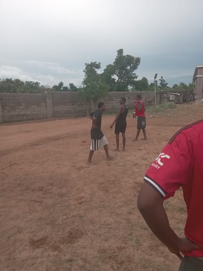 The image captures an outdoor scene in Achara Ndiagu, Nigeria. Three people are seen performing what appears to be a physical exercise or drill. They are aligned horizontally across the frame, dressed in athletic wear. The environment consists of a dirt ground with sparse vegetation, a low wall or fence made of concrete blocks, trees, and a building in the background. The sky is visible with a hazy appearance. The shot is taken at ground level.