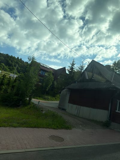 The image captures a scene in Wilkowisko, Poland, on a cloudy day. In the foreground, a red brick sidewalk runs along a paved road. To the right of the sidewalk, a grassy area slopes upwards towards buildings. A large, multi-story building with a dark red wooden exterior and a slate roof is prominently featured on the right. It has a small white-framed window visible. Further back and to the left, another building with a green facade and a red roof, equipped with solar panels, is partially visible amidst dense green trees. The background consists of a heavily wooded hillside under a sky filled with dramatic, white and gray clouds. A thin black wire stretches diagonally across the upper portion of the image.