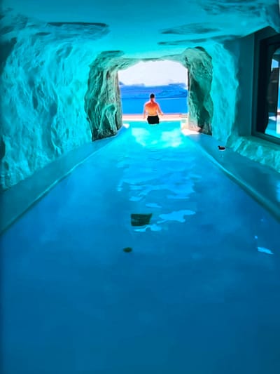 A person in black swim trunks stands facing away from the camera, their back to a surreal, cave-like swimming pool. The pool's walls are textured to resemble rock formations, with a vibrant turquoise hue. The water is a deep, rich blue, reflecting the illuminated walls.
Through an opening in the cave-like structure, a glimpse of the outside world is visible. It appears to be a bright, sunny day, with a clear blue sky and the suggestion of a beach or poolside area. The contrast between the enclosed, fantastical pool and the open, natural light outside is striking.
The person appears to be poised, perhaps about to enter the water or simply taking in the unique environment. The lighting within the pool creates an ethereal, almost otherworldly atmosphere. The overall impression is one of a luxurious and imaginative aquatic space, possibly a private pool or a high-end spa. The location context of Nijmegen, Netherlands, suggests this could be an indoor facility designed to evoke an exotic outdoor feel. There is no visible text in the image.