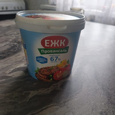 A close-up, eye-level, slightly angled shot shows a light blue plastic tub of mayonnaise on a dark gray speckled countertop. The tub has a white lid with a ribbed edge. The label on the tub features a red oval with white Cyrillic lettering that reads "ЕЖК". Below this is a green banner with white Cyrillic text that says "Провансаль". Further down, the label indicates "майонез" and "67%" in large font, with "ГОСТ 31761" below. A section of the label on the left, in a blue badge, reads "ЛЮБИМЫЙ УРАЛЬСКИЙ -ВКУС-". The bottom of the label displays a photographic image of a steak, a half tomato, a slice of lemon, and some green herbs or peas. The background is out of focus, showing what appears to be a kitchen appliance and a window with sheer curtains. The image is well-lit, with the primary subject in sharp focus. There is no indication of the specific location within the city of Юшала, Russia, beyond what can be inferred from the product's branding.