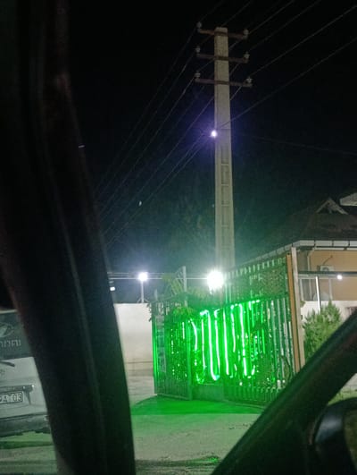 The image, taken at night from inside a vehicle, shows a street scene in Levakant, Tajikistan. In the foreground, the dark interior of the vehicle is visible, with a portion of the car's body and a window obscuring parts of the view. The background features a street with a concrete utility pole with multiple electrical wires running horizontally across the frame. To the right, a green metal fence with vertical bars is illuminated by bright green neon lights arranged in vertical stripes. Beyond the fence, a building with a sloped roof is visible, along with some greenery and a white wall. A street light with a bright white illumination is mounted on a pole in the middle ground. The license plate of a vehicle is partially visible on the left side of the image, showing the characters "9AT03".