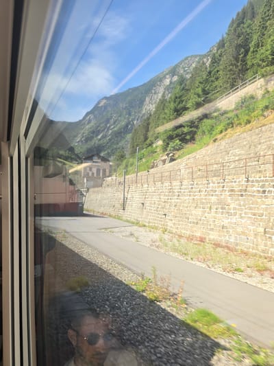 The image is a reflection in a train window, looking out at a mountainous landscape in Göschenen, Switzerland. The reflection shows a man wearing sunglasses and a light-colored shirt. Outside the window, a paved path runs alongside a stone retaining wall. Beyond the wall, a steep, tree-covered mountainside rises under a clear blue sky with a contrail. In the distance, a building with arched windows is visible. The train compartment itself is visible in the foreground, with vertical white and grey framing.