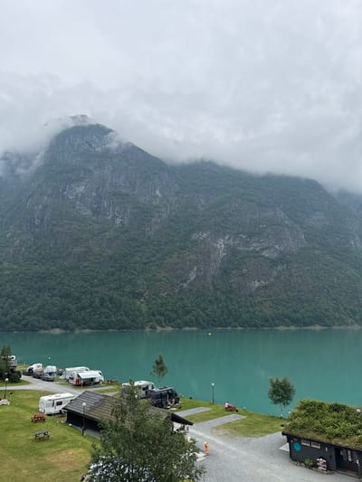 The image depicts a campsite situated by a tranquil, turquoise-colored lake, with a formidable, cloud-shrouded mountain range serving as a dramatic backdrop. The campsite is equipped with various recreational vehicles, including caravans and motorhomes, parked on a gravel and grassy terrain. Several picnic tables are scattered throughout the area, suggesting a recreational setting. A small, dark-colored building with a grass-covered roof is visible on the right side of the frame, adding a rustic charm to the scene. Two individuals are present, one seated at a picnic table near the lake and another standing near a building. The overcast sky contributes to a serene and somewhat moody atmosphere. The location is identified as Olden, Norway, a village nestled within a picturesque fjord landscape, renowned for its natural beauty and outdoor activities.