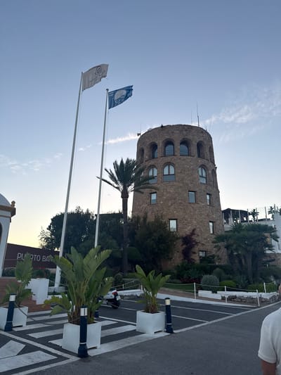 In Nueva Andalucía, Spain, a cylindrical stone tower stands prominently against a pale blue sky. Two flags, one white and one blue, are hoisted on tall poles to the left of the tower. The white flag displays a logo and text, while the blue flag features a circular graphic and dates "2025-2026". A palm tree grows to the right of the flags, partially obscuring the tower. The tower has multiple arched windows on its upper floors and smaller rectangular windows below. In the foreground, a zebra crossing leads to a paved area. Several large white planters with tropical plants are placed along the crossing and the paved area. A white scooter is parked behind some of the planters. To the far left, a white building with a decorative gold trim is partially visible, with a sign displaying "PUERTO BAJO". The environment appears to be an outdoor setting, possibly a resort or marina, with manicured landscaping and other buildings in the background. A person wearing a white shirt is partially visible on the far right of the image, their back to the camera.