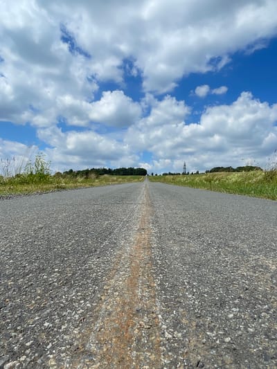 A low-angle, eye-level shot captures an asphalt road stretching towards the horizon under a bright blue sky filled with fluffy white clouds. The road appears to be in a rural setting, with dry grass and some vegetation lining its sides. In the distance, a sparse line of trees marks the edge of a field, and a cell tower is visible on the right side of the frame. The road has a distinct reddish-brown stain running down its center, suggesting it might be a former traffic lane or an area where a substance has been deposited. The overall impression is one of an open, somewhat desolate landscape. This scene is located in Schwarzenborn, Germany, as indicated by the context.
