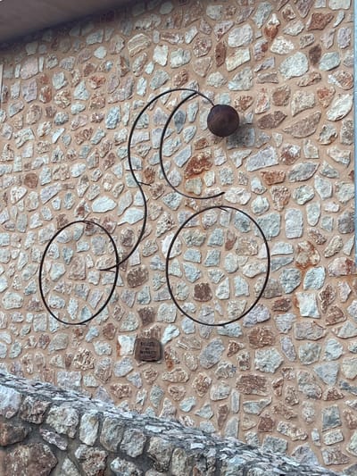 An outdoor, eye-level forensic image captures a rustic stone wall in Fornalutx, Spain, adorned with a minimalist metal bicycle sculpture. The wall is constructed from irregularly shaped, varied light-colored stones, including off-white, grey, and light brown hues, mortared together with a reddish-brown binder. The sculpture, fabricated from dark, possibly oxidized or rusted metal rods, depicts a stylized bicycle outline with two main circular wheels, a smaller third circle representing a front wheel or pedal, and a curved frame culminating in a spherical element suggesting a head or bell. Below the sculpture, centered horizontally and slightly to the left, a small, light brown rectangular plaque is embedded within the wall, bearing the inscription "CICLISTA AMIC FORNALUTX." The lower portion of the image reveals the top edge of another, darker stone wall or parapet, composed of larger, more angular grey and brown stones. The scene is static, devoid of human presence or dynamic actions.