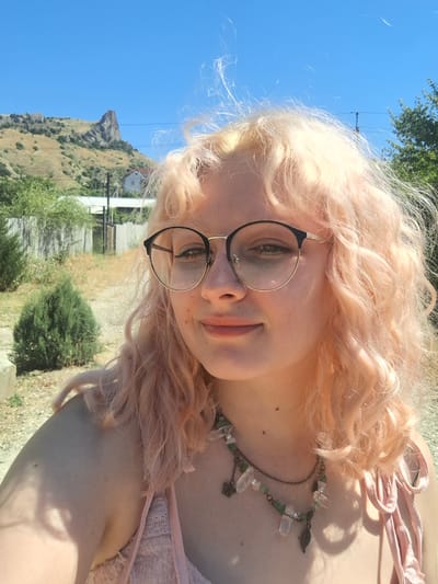 A young woman with light blonde, curly hair and round glasses is pictured in an outdoor setting. She is wearing a light pink top with a delicate lace trim and a necklace with green beads and crystal pendants. The background features a dry, grassy hillside with a distinctive rock formation at the top, a few scattered buildings, and some trees. The sky is clear and blue, indicating a sunny day. The scene appears to be in a rural or semi-rural area, potentially in or near the Щебетовский поселковый совет region of Ukraine, given the terrain and vegetation.