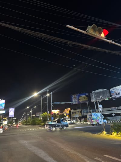 This is a nighttime, outdoor, eye-level, medium shot of a street scene in Denpasar Barat, Indonesia. The image is heavily rotated, appearing to be taken from a moving vehicle. The primary subject is a traffic light displaying a red light, indicating a stop. Surrounding the traffic light are numerous power lines, creating a sense of urban infrastructure.
The scene depicts a street at night, with the asphalt road visible and illuminated by streetlights and vehicle headlights. Several vehicles are present, including a dark-colored car in the foreground and a white van with colorful graffiti in the middle ground. The van appears to be parked or stopped. In the background, there are buildings and trees, suggesting an urban or suburban environment. Several billboards and advertisements are visible along the roadside, some with text.
Notable details include the bright illumination from streetlights, the dark sky indicating nighttime, and the general atmosphere of a busy street after dark. The presence of traffic lights and vehicles suggests a functional roadway. The overall mood is that of a typical urban night, possibly with some rain or wetness on the road due to the reflections of light.
Visible text includes partial words on billboards. One billboard has text that appears to be in Indonesian, possibly related to advertising or public announcements. The overall impression is a slice of life in a bustling Indonesian city at night.
