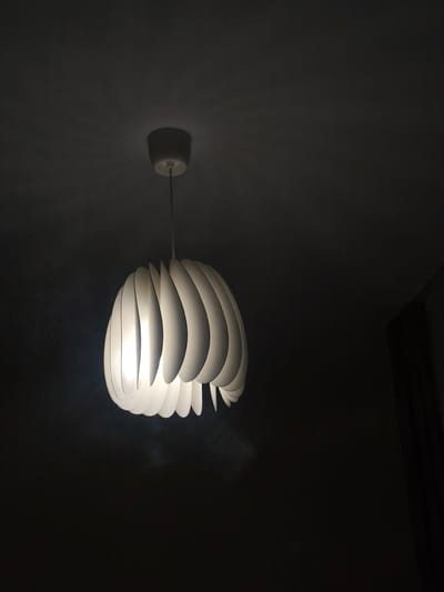 A lit, white, decorative lamp hangs from a white base attached to a dark ceiling. The lamp consists of numerous curved, overlapping white segments arranged in a spherical shape. The surrounding environment is dark, with a dark wall visible in the lower right corner. The location is not discernible from the image. There is no evidence in the image to confirm the location in Mosomishte, Bulgaria. There are no people present in the image.