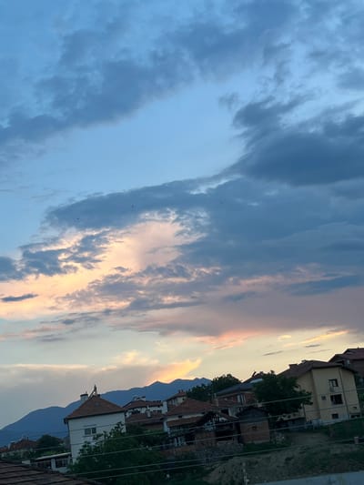 This is an outdoor, eye-level, medium shot photograph taken in Ognyanovo, Bulgaria. The image depicts a residential area with several houses nestled against a backdrop of rolling hills and a dusky sky. The sky is a mix of blues and grays, with streaks of pale pink and orange hinting at the setting sun. Dark, wispy clouds dominate the upper portion of the frame, with lighter, more translucent clouds scattered lower down. A few dark birds are visible in flight against the sky. The houses are varied in style and color, with terracotta tile roofs being a common feature. Some houses appear well-maintained, while others show signs of disrepair. Green trees and foliage are interspersed among the buildings. In the distance, dark mountain silhouettes rise against the horizon, topped by a single, illuminated tower. The overall impression is one of a quiet, rural evening scene in a Bulgarian village.