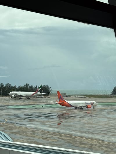 The image depicts an airport tarmac on a cloudy day, possibly during or after a rain shower, as evidenced by the wet surfaces. In the foreground, the tarmac is visible with yellow and white markings, reflecting the overcast sky.

Two commercial airplanes are present on the tarmac. The aircraft on the left is an Emirates airline plane, identifiable by its distinct livery and logo. It is parked on the tarmac, with its engines visible. The aircraft on the right is an Akasa Air plane, also clearly marked with its branding. This plane appears to be either taxiing or has just landed, as it is positioned on a taxiway or runway.

Behind the planes, a line of trees separates the tarmac from the sea in the distance. The sky is predominantly gray and cloudy, creating a diffused light over the scene. The overall impression is of a busy airport environment, with the presence of two large passenger jets indicating ongoing air traffic. The location is identified as Ban Yid, Thailand.