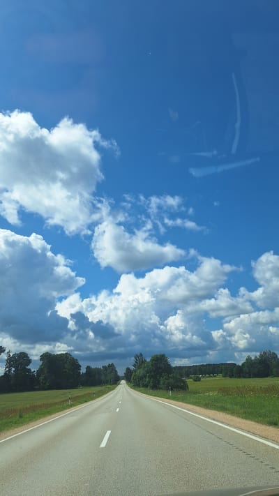 The image shows an asphalt road stretching into the distance, bordered by green fields and trees on either side. The sky is a vibrant blue with scattered white clouds, some of which are large and fluffy. The road has white lane markings. The scene appears to be a rural landscape, possibly in the city of Glāžšķūnis, Latvia, based on the provided context. There are no people or vehicles visible in the image. The overall impression is one of a clear, sunny day on an open road.