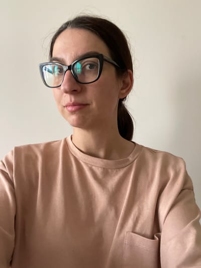 A woman with dark hair pulled back and wearing black-rimmed glasses is pictured against a plain, light-colored wall. She is wearing a light pink t-shirt with a breast pocket. The reflection in her glasses suggests an interior setting. The photograph’s orientation is tilted. There is no information about Shishmantsi, Bulgaria, in the image.