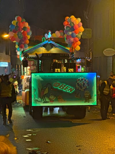 The image depicts a nighttime street scene in Wasserbillig, Luxembourg, featuring a decorated parade float. The street, composed of wet asphalt, reflects ambient light and shows scattered debris.

The central subject is a large, elaborate parade float, likely built on a truck or tractor chassis, viewed from the rear. The float's upper section features a vibrant blue and yellow house-like structure, topped with a red and blue rooster weather vane. Two substantial clusters of multicolored balloons (red, orange, yellow, green, blue, purple) are affixed to either side of the house, evocative of the Disney/Pixar movie "Up." Green neon-style lighting outlines elements of the float's structure.

The rear of the float features a large, rectangular, illuminated display panel. This panel showcases a cartoon scene from "Up," depicting characters Kevin the bird, Russell, Carl Fredricksen, and Dug the dog, alongside a waterfall. The German word "OBEN" (meaning "UP") is prominently displayed in illuminated letters. German text above reads: "Das Abenteuer beginnt, es wird hoch geflogen, die Laasa Allstars präsentieren." (The adventure begins, it flies high, the Laasa Allstars present.)

Numerous individuals are present. At least one person is partially visible within the house structure on the float. Spectators line both sides of the street, partially obscured by the float and foreground elements. Two individuals wearing yellow reflective vests, possibly event staff or security, are visible on the left and right sides of the float.

The surrounding environment includes multi-story buildings lining the street. A white sign with blue text "Staudt" is visible on the left. On the right, a brown sign with "Peinture Bornsmark" and another sign above it are visible, indicating commercial establishments. A blue and white pedestrian crossing sign is partially visible above the float on the right. Artificial light sources from streetlights and building interiors illuminate the scene in addition to the float's own lighting.