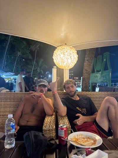 The image captures two adult males seated outdoors at night under a white canopy with a spherical, textured light fixture, located in Cairu, Brazil. The man on the left is shirtless, wearing dark shorts, a grey cap, and glasses, with a dark mustache. He has a red wristband on his left wrist and a visible tattoo on his right arm. His left hand is raised in a gesture with fingers spread, while his right hand holds a lit cigarette. On the dark wooden table to his left are a plastic water bottle with a blue label and a black pouch. The man on the right has light-colored curly hair and a beard, wearing a dark t-shirt and red and dark shorts. He has a red wristband on his left wrist and a tattoo on his left forearm. His left hand is raised in a "shaka" gesture, and his right hand holds a cigarette. On the table to his right are a red Coca-Cola Zero can, a white plate with orange food residue and a fork, and a small white packet. In the background, palm trees are visible against a dark sky, along with several other individuals and indistinct structures, including a green, possibly sculptural, element.
