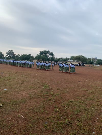 The scene depicts an outdoor gathering in Kubi A, Nigeria, taking place on a large, reddish-brown dirt field sparsely covered with green grass. The sky is uniformly overcast with light grey clouds. In the background, numerous green trees line the horizon, interspersed with low-lying buildings, a prominent water tower with a white tank on a metallic frame, and a tall pole equipped with a solar panel and light fixture. Several vehicles are parked further in the distant background to the right.
A large group of approximately 100-150 individuals, primarily young adults, are organized into multiple parallel lines across the field. They are uniformly dressed in what appears to be the National Youth Service Corps (NYSC) attire, consisting of white short-sleeved shirts, green trousers, white caps, and brown footwear. The individuals are positioned in structured formations, with the longest line extending from the mid-ground on the left towards the background, and another distinct formation of shorter lines positioned closer to the foreground on the right. Their posture suggests they are either standing at attention or engaged in a drill. One individual, distinctively dressed in a camouflage uniform, stands between the two main formations, possibly supervising or instructing. There are no clear discernible interactions between individuals, as they maintain their disciplined formations.